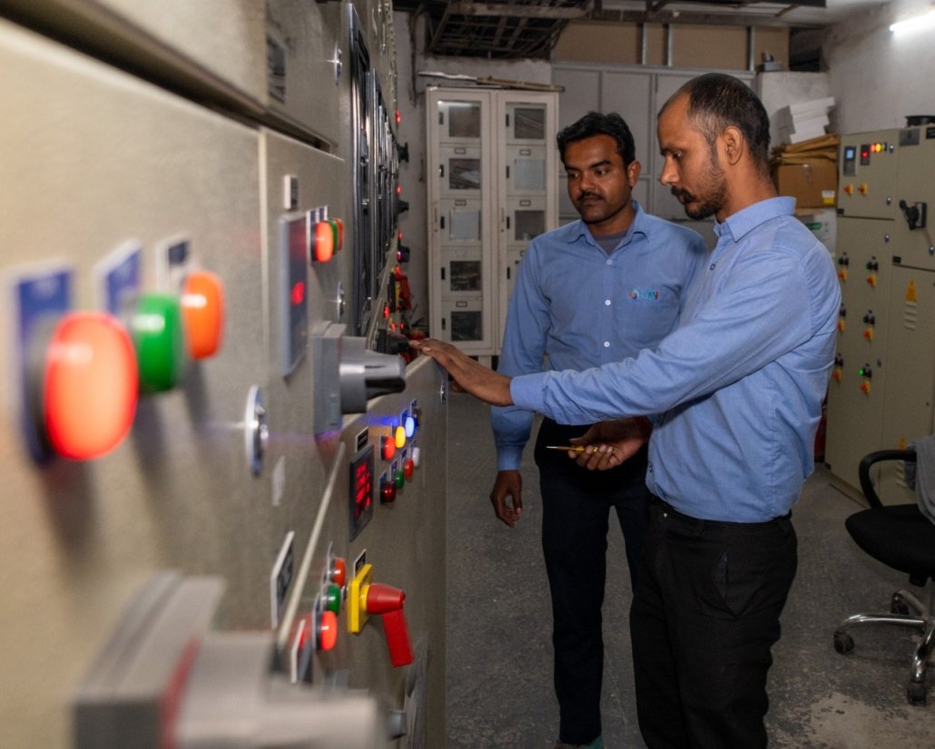 Two technicians from CAS FACILITIES wearing blue shirts operate and monitor controls on a large industrial control panel with illuminated buttons and switches inside a control room.