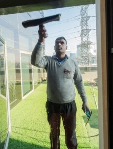 A Cas Facilities worker cleaning an exterior glass window using a squeegee during daylight.